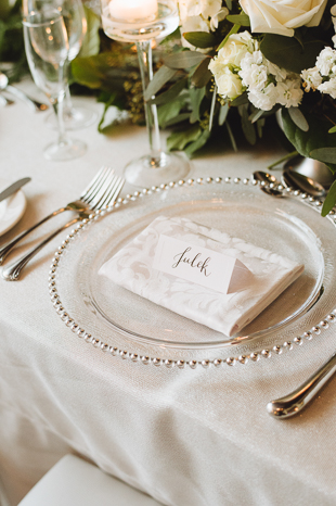 Liberty Grand Wedding Photography. Wedding details photography of guest table settings. Clear glass plates with white place setting napkins and florals.